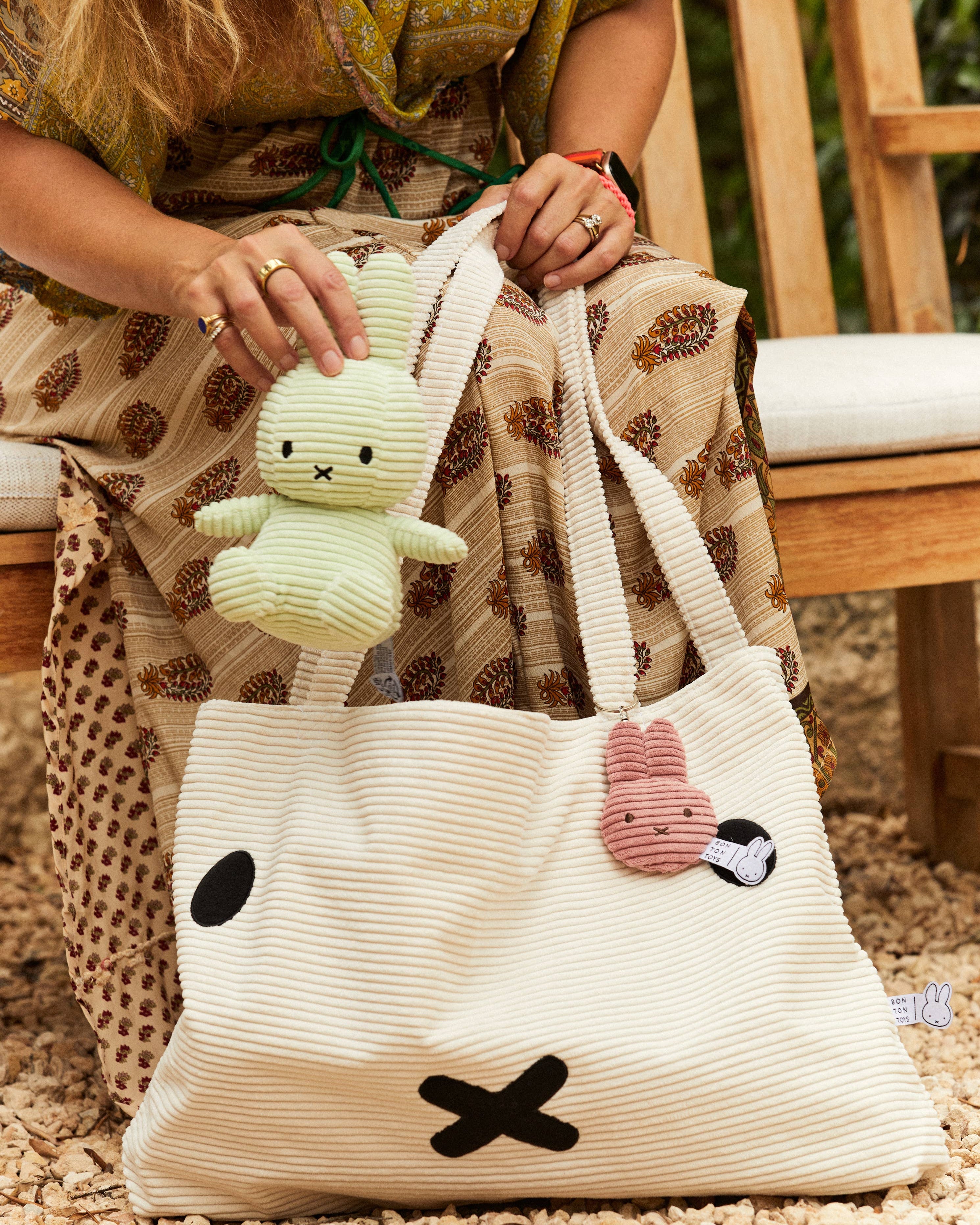Person holding a white tote bag with cartoon faces and a small green plush Miffy toy, sitting on a wooden bench.