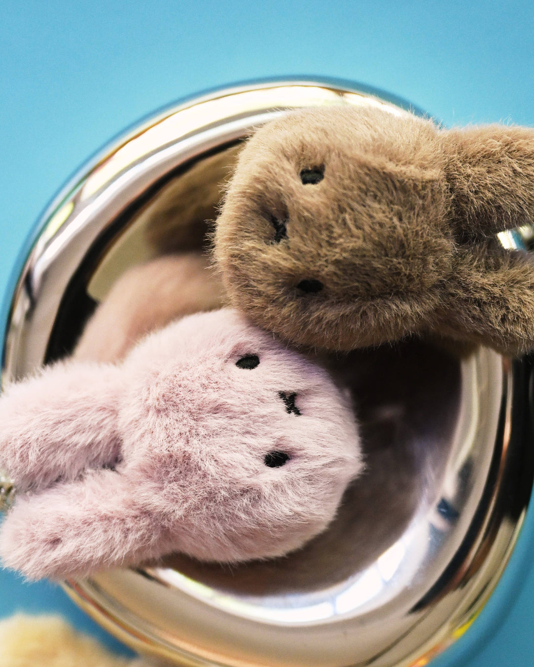 Two fluffy Miffy keychains, one brown and one pink, on a metallic surface with a blue background.